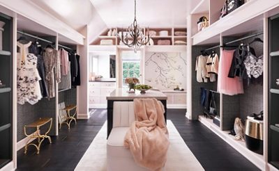 Large sunlit walk-in closet with pastel pink walls, clothing racks, elegant chandelier, and mural of a branch on one wall.