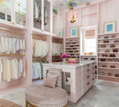 Pastel pink walk-in closet with high wallpapered ceiling, gold accents, marble-topped island, and neatly displayed shoes and clothing.