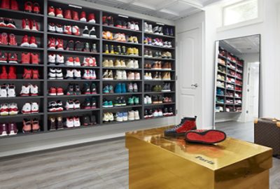 Custom shoe room with dark shelves full of colorful sneakers, white walls, large leaning mirror, and a gold bench in foreground.