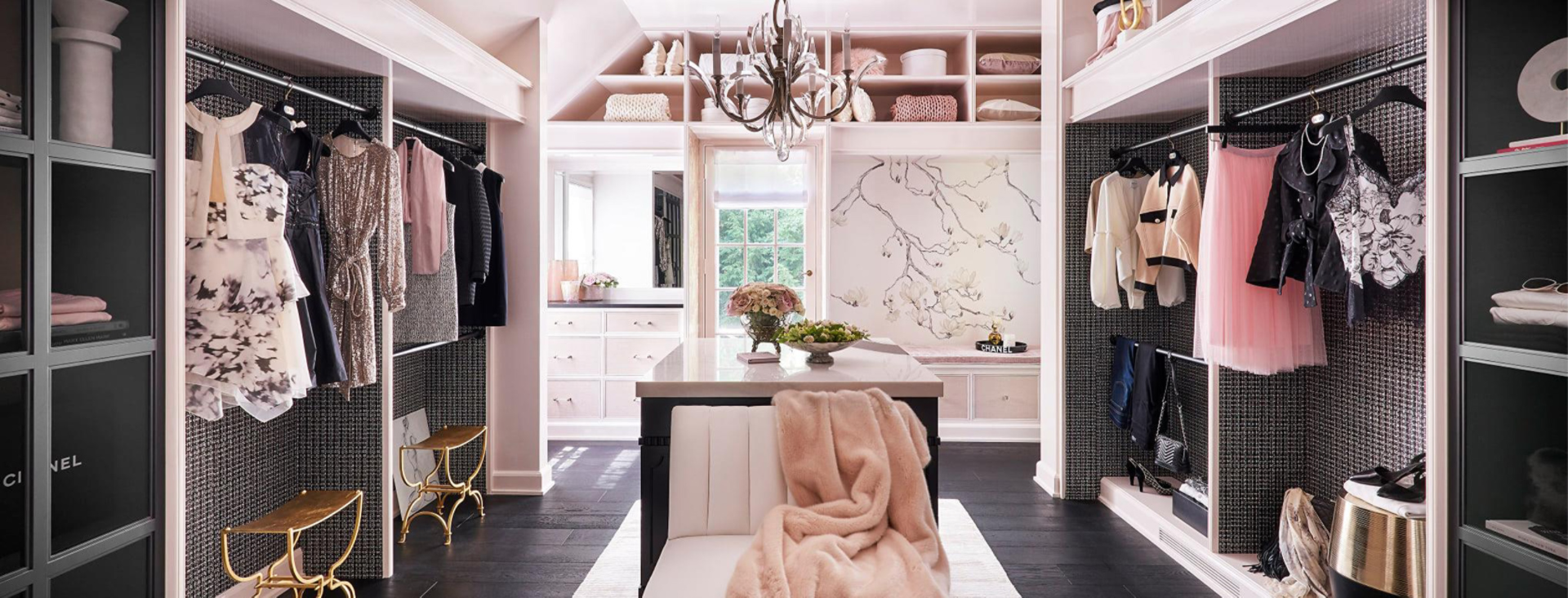 Large sunlit walk-in closet with pastel pink walls, clothing racks, elegant chandelier, and mural of a branch on one wall.