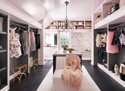 Large sunlit walk-in closet with pastel pink walls, clothing racks, elegant chandelier, and mural of a branch on one wall.