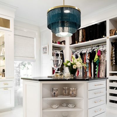 Closer look at walk-in closet center island with black top, dark teal fringe chandelier, hanging clothes and window behind.