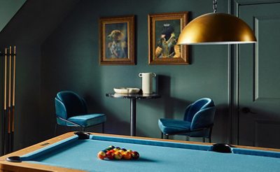 A stylish residential recreation room color-drenched in Pewter Green by Sherwin-Williams, featuring a sofa, billiard table, and antique brass decorative elements.