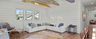 A modern living room with a white sectional sofa, neon "stay + play" sign, and surfboards on the ceiling, with walls and trim painted Sherwin-Williams Pure White.