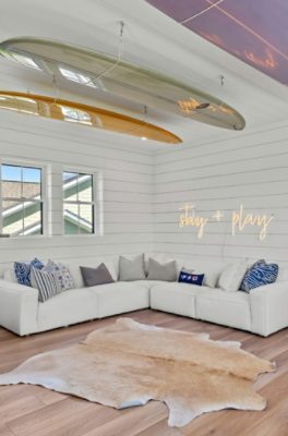 A modern living room with a white sectional sofa, neon "stay + play" sign, and surfboards on the ceiling, with walls and trim painted Sherwin-Williams Pure White.