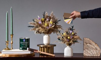 Gray wall in the background of a wooden table with 2 candlesticks, a can of Emerald paint, paintbrush, 2 vases of East Olivia floral bouquets.