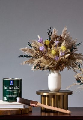 Gray wall in the background of a wooden table with a can of Emerald paint, paintbrush, a vase of East Olivia floral bouquets.
