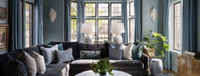 Living room with a dark gray sectional sofa, assorted pillows, marble coffee table with greenery, and tall windows with blue curtains.