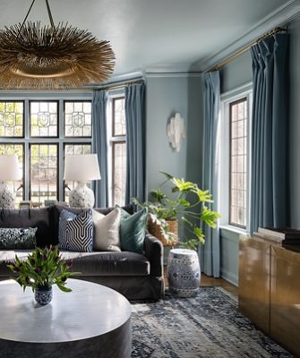 Living room with a dark gray sectional sofa, assorted pillows, marble coffee table with greenery, and tall windows with blue curtains. 