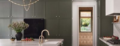 Modern kitchen with dark Roycroft Bronze Green cabinets, marble countertop, gold faucet, and a window view of a garden.
