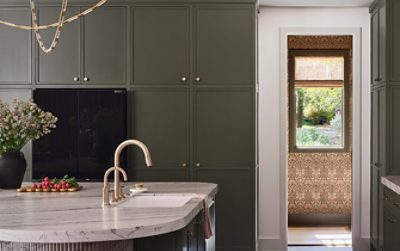 Modern kitchen with dark Roycroft Bronze Green cabinets, marble countertop, gold faucet, and a window view of a garden.