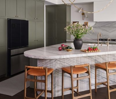 Modern kitchen with marble backsplash and island topped with vase of flowers and fresh vegetables, and Roycroft Bronze Green perimeter cabinets. 