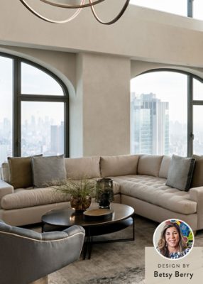 Modern living room with neutral color scheme, large windows, a beige sectional sofa, a dining table, and a marble kitchen island, with a headshot of designer Betsy Berry in the corner of the image.