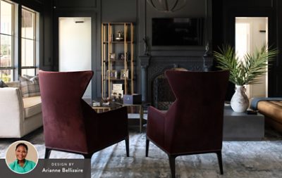 Luxurious living room with charcoal walls, vintage fireplace, burgundy armchairs, beige sofa, and a palm frond vase on a coffee table, with a headshot of designer Arianne Bellizaire in the corner of the image. 