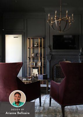 Luxurious living room with charcoal walls, vintage fireplace, burgundy armchairs, beige sofa, and a palm frond vase on a coffee table, with a headshot of designer Arianne Bellizaire in the corner of the image. 