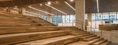 Wide wooden stairway inside a modern office space with stair steps at right and tiered stadium bench seating at left.