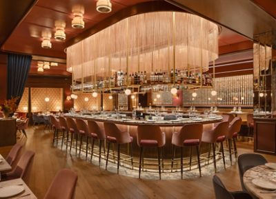 A luxurious restaurant bar with a rounded counter, pink bar stools, and a golden overhead bottle rack and fringed lighting fixture. 