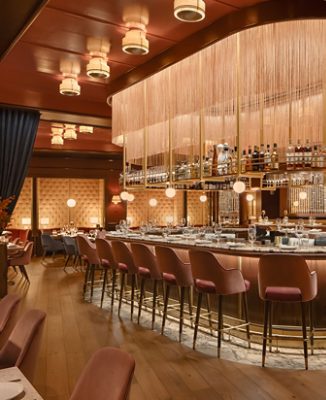A luxurious restaurant bar with a rounded counter, pink bar stools, and a golden overhead bottle rack and fringed lighting fixture. 