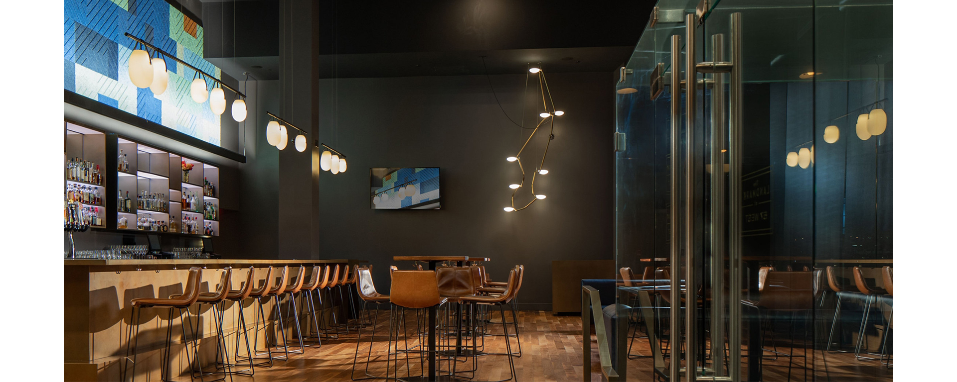 Moody dark interior of restaurant with wood floors, high ceilings, modern lighting, and leather chairs at wood tables and bar.