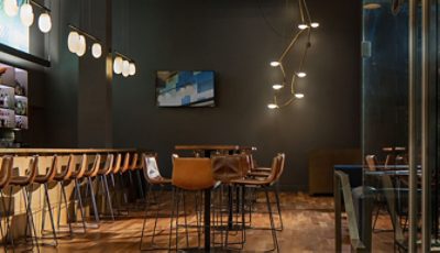 Moody dark interior of restaurant with wood floors, high ceilings, modern lighting, and leather chairs at wood tables and bar.