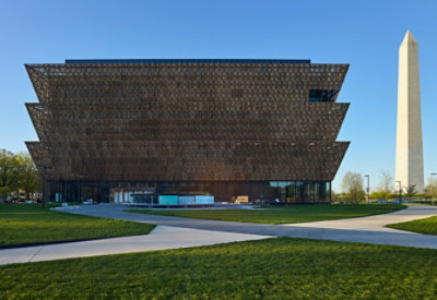 Exterior of the National Museum of African American History and Culture in front of blue sky and with the Washington Monument visible at right.