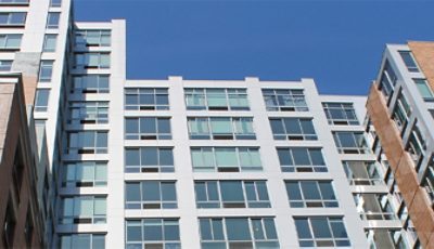 Exterior shot looking up at a city apartment building with brick and metal exterior, wide windows, and clear blue sky above.