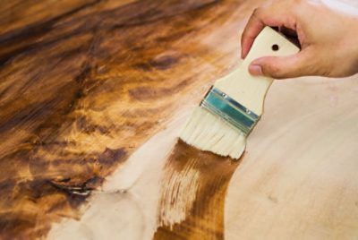 Close-up of a person's hand applying wood stain with a stain brush to a wooden surface.