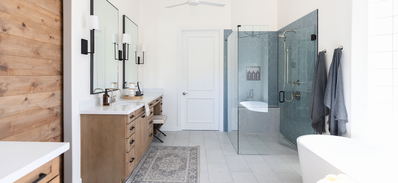 Accessible primary bathroom with glass-walled shower, natural wood double vanities and a freestanding modern bathtub.