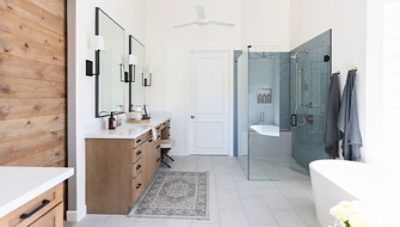 Accessible primary bathroom with glass-walled shower, natural wood double vanities and a freestanding modern bathtub.