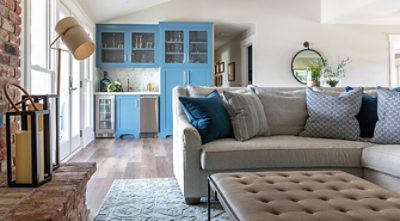 Living room with neutral sectional and brick fireplace in foreground, wet bar with blue cabinetry in the background.