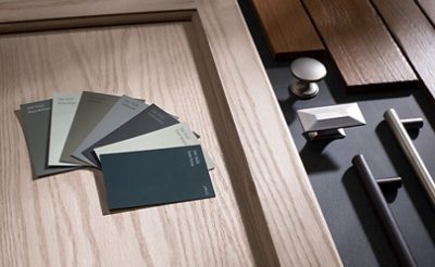 Closeup of blue, gray, and green paint chips on top of light wood cabinet door next to a variety of metal drawer pulls on a dark surface.