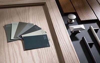 Closeup of blue, gray, and green paint chips on top of light wood cabinet door next to a variety of metal drawer pulls on a dark surface.