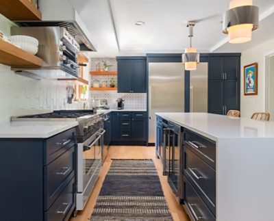 Modern kitchen with open wood shelving, modern stainless steel appliances and lighting, sleek white island, and dark blue cabinetry.
