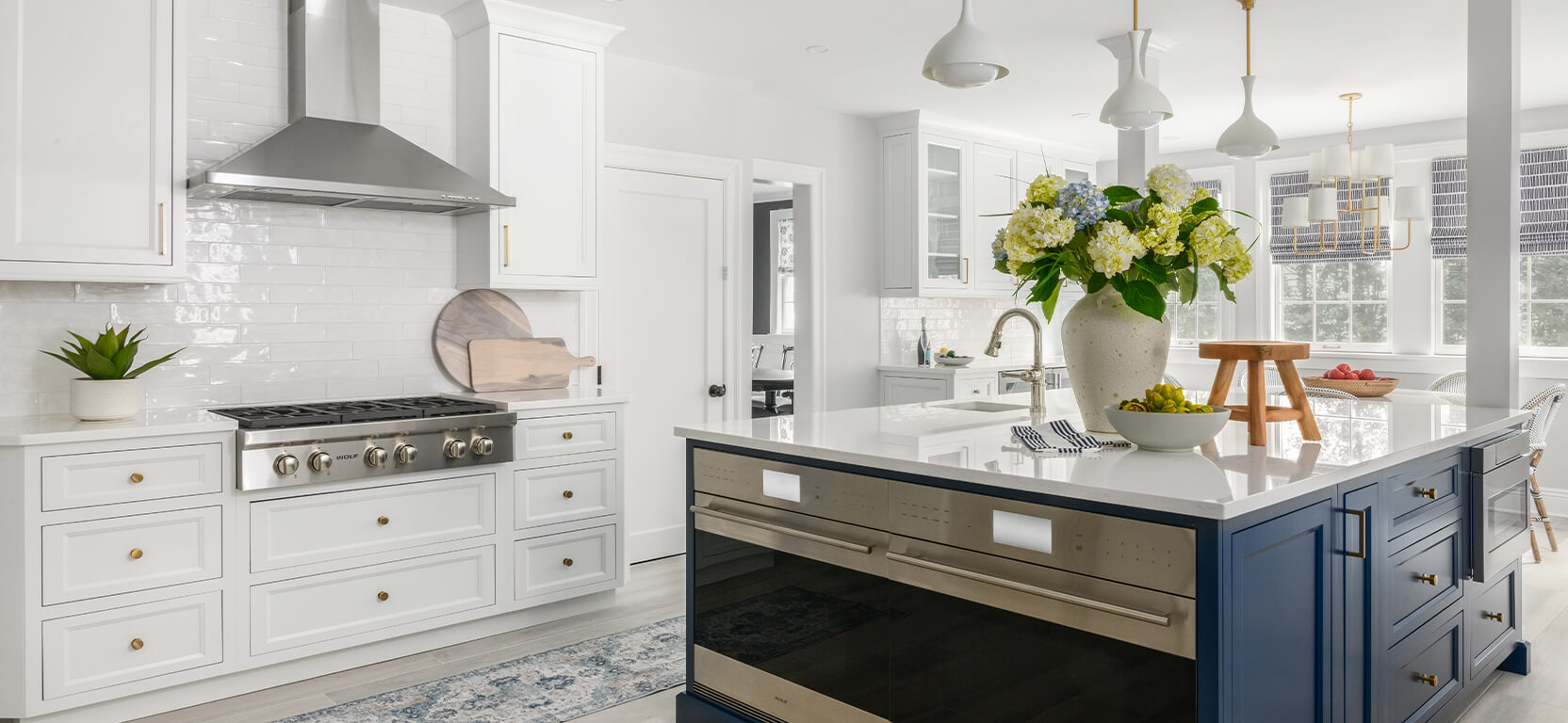 Sunlit white kitchen with double ovens in the center kitchen island, which is painted a deep contrasting shade of navy blue.