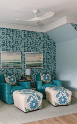 A sitting area on a second-floor landing with patterned blue wallpaper, two teal armchairs with floral footrests, and landscape art prints featuring Watery and Pure White paint colors by Sherwin-Williams.