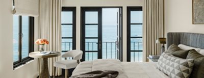 A Cheviot-painted bedroom with a view of the ocean through large black-framed windows, furnished with a bed, wooden table, and a chair.