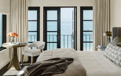A Cheviot-painted bedroom with a view of the ocean through large black-framed windows, furnished with a bed, wooden table, and a chair.