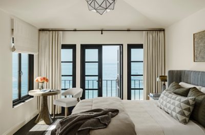 A Cheviot-painted bedroom with a view of the ocean through large black-framed windows, furnished with a bed, wooden table, and a chair. 
