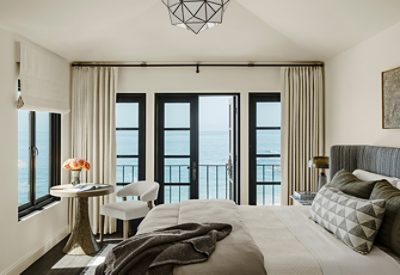 A Cheviot-painted bedroom with a view of the ocean through large black-framed windows, furnished with a bed, wooden table, and a chair. 