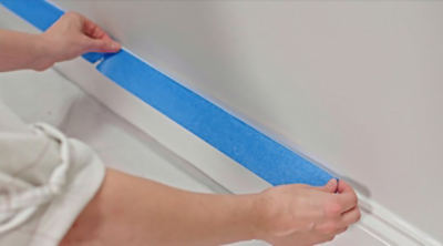 A person using blue painter's tape to mask wall trim before painting