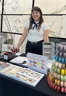 Woman in a white shirt and dark pants smiling while standing at a table with an array of Sherwin-Williams color tools and tiered display of colorful macarons.