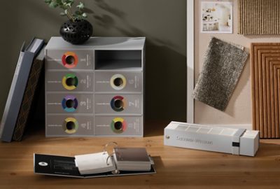 Gray box of color sample binders, small ringed binder of samples, and color fan deck on a wooden desktop with fabric samples and photos on a pinboard and a potted plant.