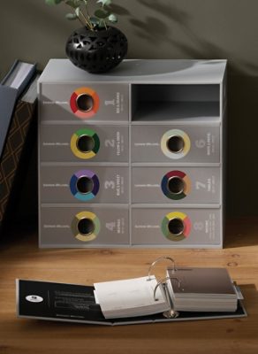Gray box of color sample binders, small ringed binder of samples, and color fan deck on a wooden desktop with fabric samples and photos on a pinboard and a potted plant.