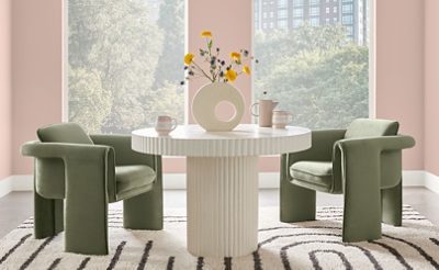 Dining area for two with modern fluted white pedestal table, plush sage green velvet armchairs in front of large windows and a backdrop of walls painted Sashay Sand by Sherwin-Williams.