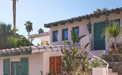 Exterior of the Mesa Bungalows project, a Spanish adobe style home painted Charming by Sherwin-Williams with green doors and windows, surrounded by palm trees.