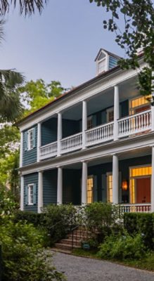 A two-story blue house with white trim, featuring front porches supported by columns, surrounded by greenery.