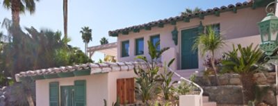 Exterior of the Mesa Bungalows project, a Spanish adobe style home painted Charming by Sherwin-Williams with green doors and windows, surrounded by palm trees.
