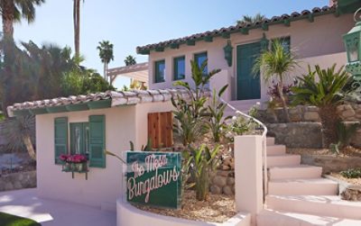 Exterior of the Mesa Bungalows project, a Spanish adobe style home painted Charming by Sherwin-Williams with green doors and windows, surrounded by palm trees.