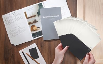 Hands holding paint chip sheen samples fanned out over the Sherwin-Williams Guide to Gloss and Sheen laying open on a wood surface.