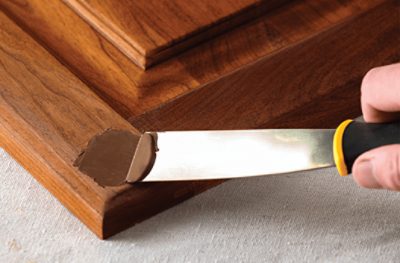 A person fixing a damaged corner of a piece of wood furniture using a putty knife.